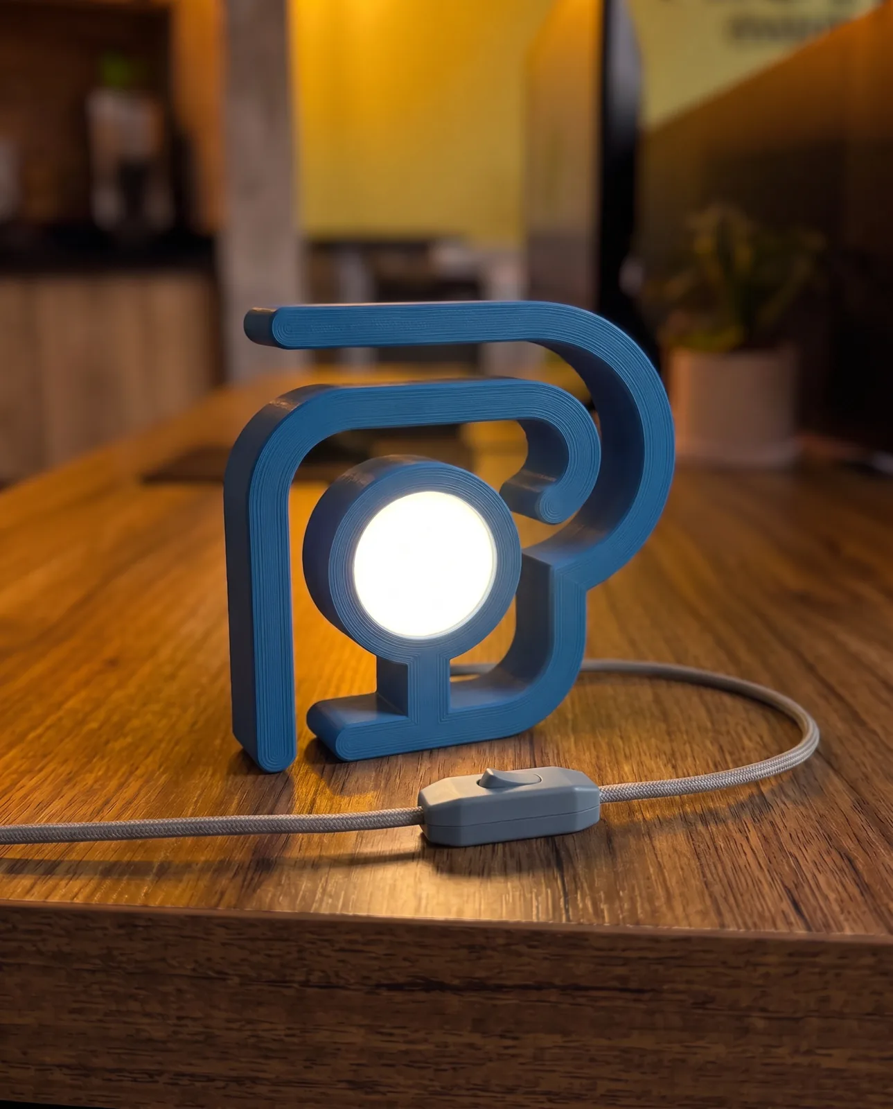 Luminaria azul impressa em 3D com o simbolo da marca, acesa sobre bancada de madeira